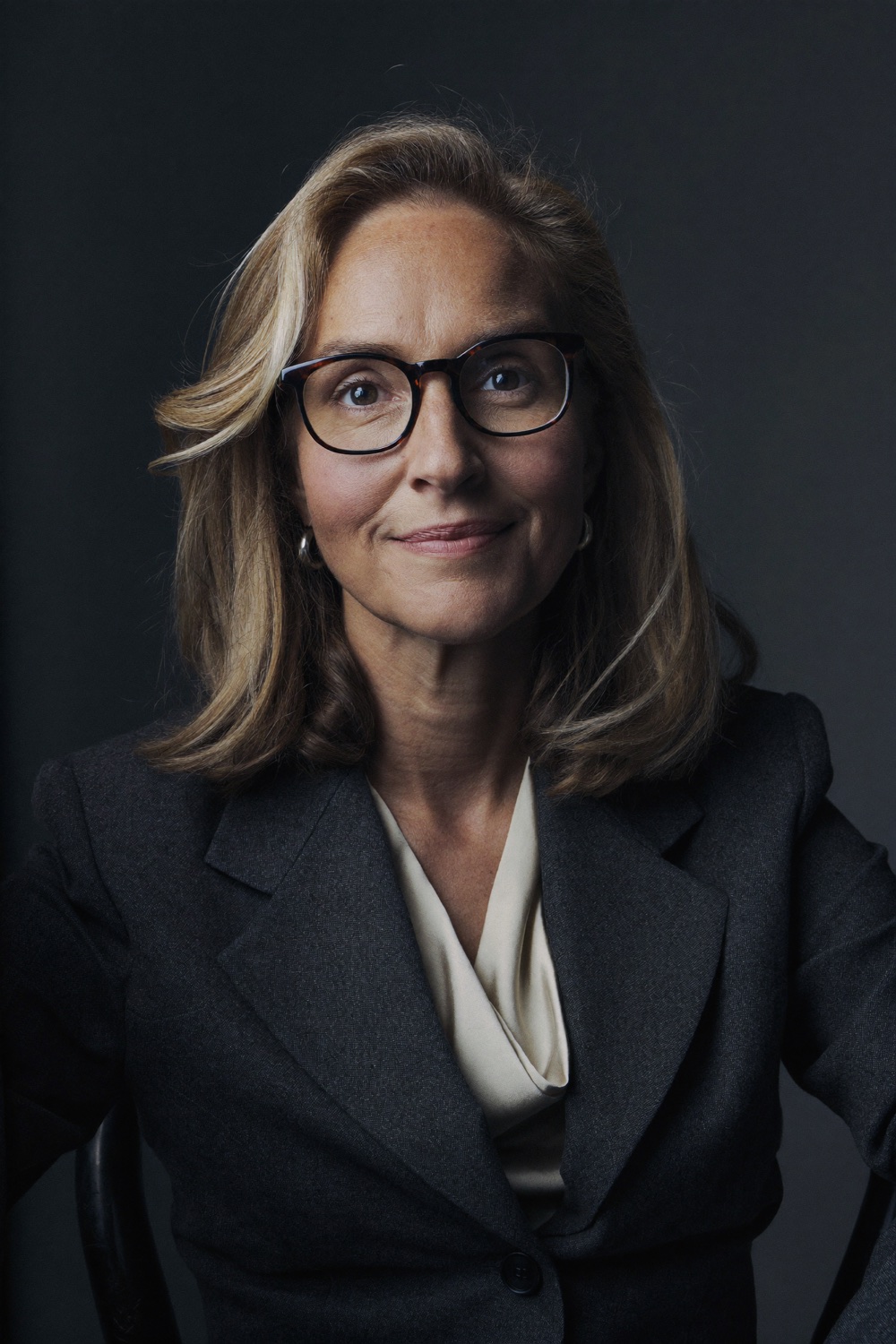 A woman with shoulder-length blonde hair and dark-rimmed glasses, in a charcoal blazer and cream blouse